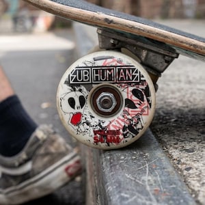 May include: Close-up of a skateboard wheel with the text "SUB HUMANS" and a skull graphic. The wheel is white with black and red accents. The skateboard is resting on a concrete ledge.