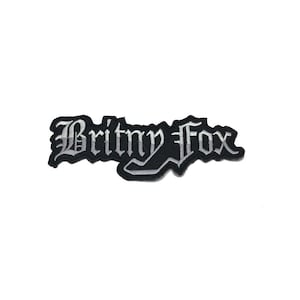 May include: A black and white embroidered patch with the text "Britny Fox" in a gothic font. The patch has a black border and is likely designed to be sewn or ironed onto clothing or accessories.