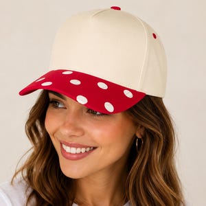 May include: A five-panel baseball cap featuring a solid cream-coloured crown and a vibrant red brim decorated with white polka dots. The cap includes a small red button at the top centre and a structured design with a curved visor.