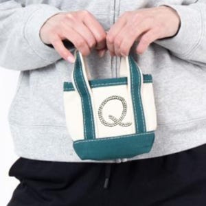 May include: A small tote bag with a white body, teal base and handles, and a teal border. The bag features a large, embellished letter "Q" on the front. The bag is held by a person wearing a gray zip-up hoodie.