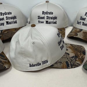 Può includere: Una collezione di sei cappellini da baseball con corona bianca e visiera mimetica. Ogni cappellino presenta un testo ricamato in nero, incluse frasi come Hydrate Shoot Straight Stay Married, Hydrate Shoot Straight Found The One e Asheville 2026.
