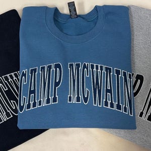 Może przedstawiać: Trzy bluzy z okrągłym dekoltem w kolorze czarnym, niebieskim i szarym melanżowym. Każda odzież posiada napis "CAMP MCWAIN" wydrukowany pogrubioną, łukowatą czcionką w stylu uniwersyteckim na klatce piersiowej. Czarne i niebieskie koszule mają białe litery, a szara koszula ma czarne litery.