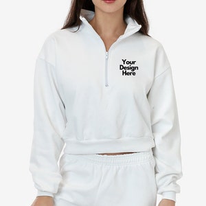 Puede incluir: Sudadera blanca recortada con cierre de cremallera y mangas largas. La sudadera tiene el texto "Your Design Here" impreso en la parte delantera.