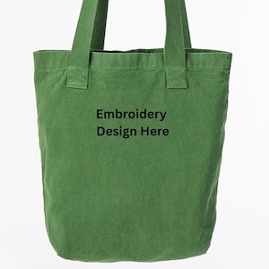 May include: A green canvas tote bag with long handles is held up by a person. The bag has the text "Embroidery Design Here" printed on the front. The bag is a solid green color.