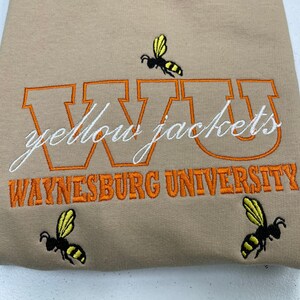 May include: Tan sweatshirt with orange embroidered lettering that reads "WVU Yellow Jackets Waynesburg University". Three yellow and black bee illustrations are embroidered on the sweatshirt.