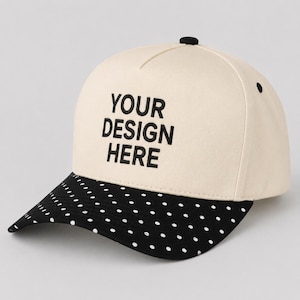 May include: A cream-colored baseball cap featuring the black embroidered text YOUR DESIGN HERE on the front panel. The cap has a black brim decorated with a repeating pattern of small white polka dots and a black button on the top crown.