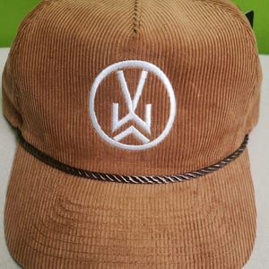 Puede incluir: Una gorra de b&eacute;isbol de pana marr&oacute;n con un logotipo bordado en blanco de un c&iacute;rculo con una "W" estilizada en su interior. La gorra tiene un ribete de cuerda marr&oacute;n alrededor de la visera.