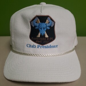 Puede incluir: Gorra de b&eacute;isbol de pana blanca con un logotipo bordado de un toro azul y un hacha en la parte delantera. El logotipo dice "Lumberjacks Life" y "Est. 2022". El texto "Club President" est&aacute; bordado debajo del logotipo.