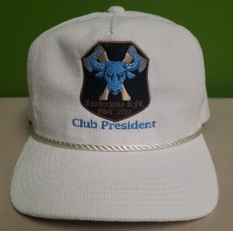 Puede incluir: Gorra de b&eacute;isbol de pana blanca con un logotipo bordado de un toro azul y un hacha en la parte delantera. El logotipo dice "Lumberjacks Life" y "Est. 2022". El texto "Club President" est&aacute; bordado debajo del logotipo.