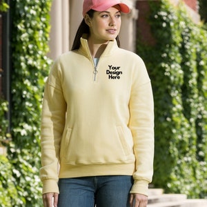 Peut inclure: Un sweat-shirt jaune pâle avec un col montant zippé et une poche avant. Le texte "Your Design Here" est imprimé sur la poitrine. Le sweat-shirt a des manches longues et une coupe décontractée. Le modèle porte une casquette de baseball rose.