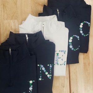 May include: Four navy blue and white quarter-zip sweatshirts with floral embroidery spelling out the word "J-N-E-C".