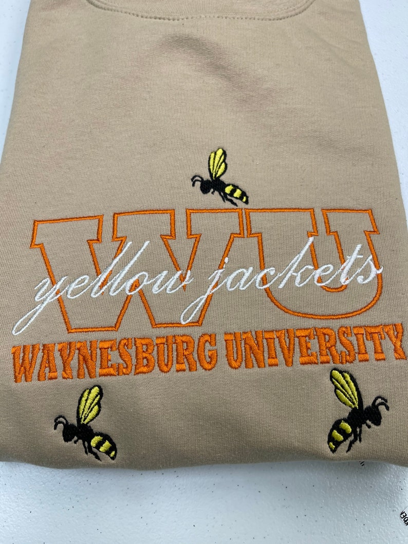May include: Tan sweatshirt with orange embroidered lettering that reads "WVU Yellow Jackets Waynesburg University". Three yellow and black bee illustrations are embroidered on the sweatshirt.