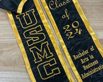 May include: Black graduation stole with gold trim and the text "USMC" on one side and "Class of 2024 Bachelor of Arts in Business Administration" on the other side. The stole also features a red patch with the USMC emblem and a small American flag.