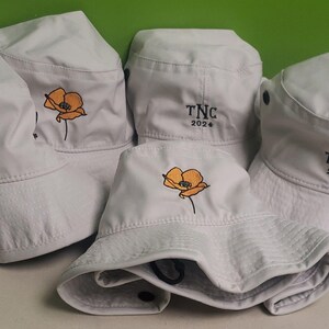May include: Four white bucket hats with different embroidered designs. One hat has a poppy flower, another has a poppy flower and the text "TNC 2024", and the other two hats have the text "TNC 2024".
