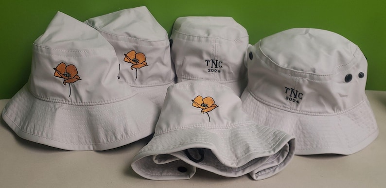 May include: Four white bucket hats with different embroidered designs. One hat has a poppy flower, another has a poppy flower and the text "TNC 2024", and the other two hats have the text "TNC 2024".