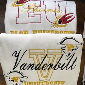 May include: Two white sweatshirts with embroidered logos. The top sweatshirt has a red and gold embroidered logo that says "Phoenix University". The bottom sweatshirt has a black and gold embroidered logo that says "Vanderbilt University".