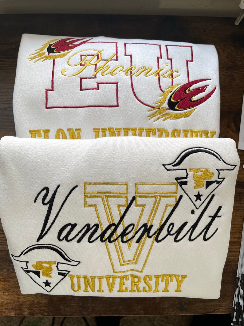 May include: Two white sweatshirts with embroidered logos. The top sweatshirt has a red and gold embroidered logo that says "Phoenix University". The bottom sweatshirt has a black and gold embroidered logo that says "Vanderbilt University".