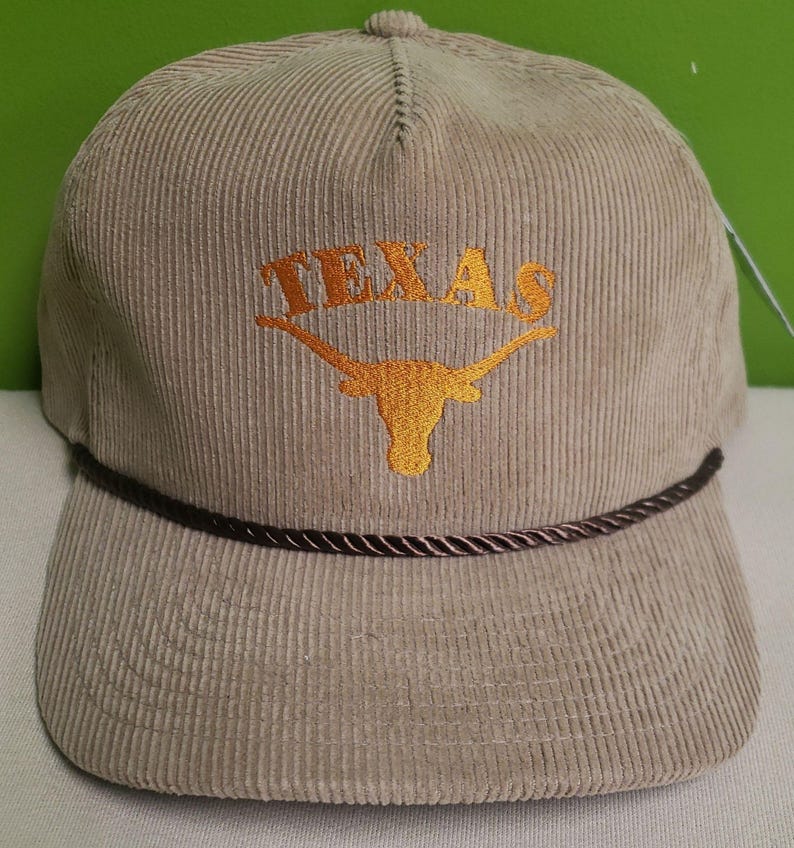 Puede incluir: Una gorra de b&eacute;isbol de pana beige con ribete de cuerda marr&oacute;n. La gorra tiene un logotipo bordado de Texas Longhorn naranja con la palabra "TEXAS" encima.