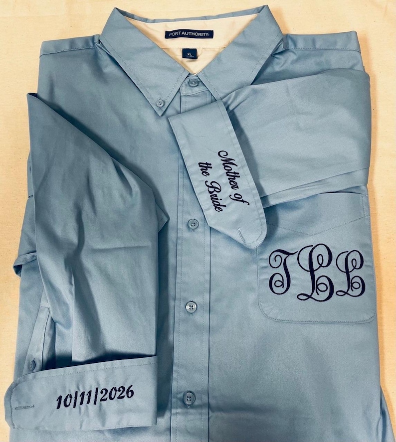 K&ouml;nnte beinhalten: Ein hellblaues Hemd mit Knopfleiste, Port Authority-Etikett und XL-Gr&ouml;&szlig;enangabe. Das Hemd ist dunkelblau bestickt: auf der Brusttasche mit dem Monogramm JLL, auf einer Manschette mit Mother of the Bride und auf der anderen mit 10/11/2026.