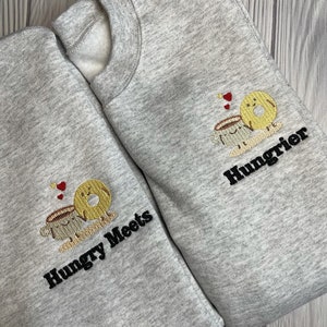 Pode incluir: Duas sweatshirts cinzas com desenhos bordados. Uma sweatshirt tem uma xícara de café e um donut com o texto "Hungry Meets" e a outra tem uma xícara de café e um donut com o texto "Hungrier".