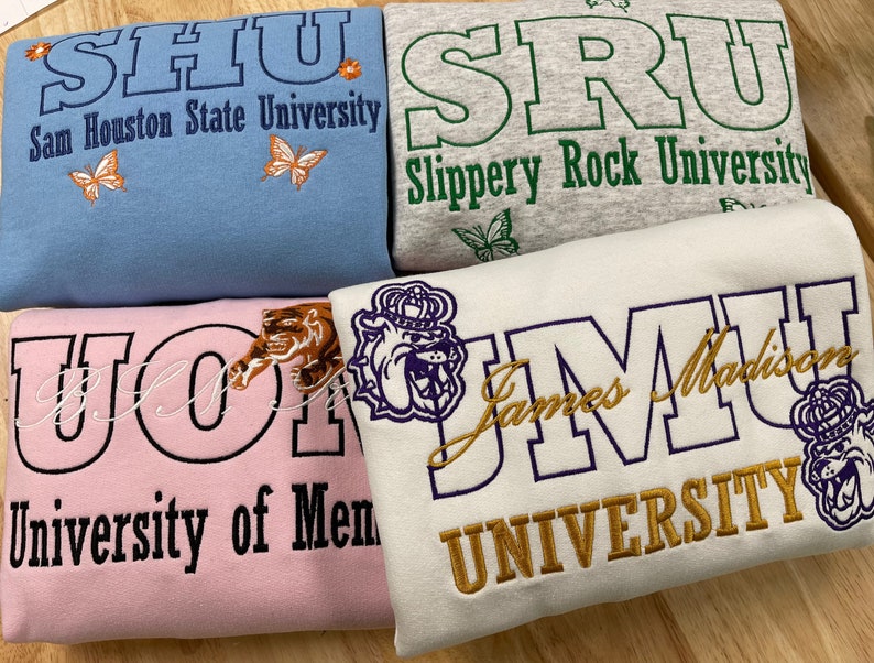 May include: Four different sweatshirts with embroidered logos for various universities. The sweatshirts are in pink, light blue, gray, and white. The logos are in black, purple, green, and gold embroidery. The logos include the names of the universities: Sam Houston State University, Slippery Rock University, University of Memphis, and James Madison University.
