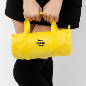 May include: A bright yellow duffel bag with matching yellow handles and straps. The bag has the text "Your Design Here" printed in black. The bag is cylindrical in shape and appears to be made of a lightweight material.