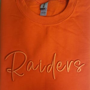 May include: Orange sweatshirt with the word "Raiders" embroidered in a cursive font.