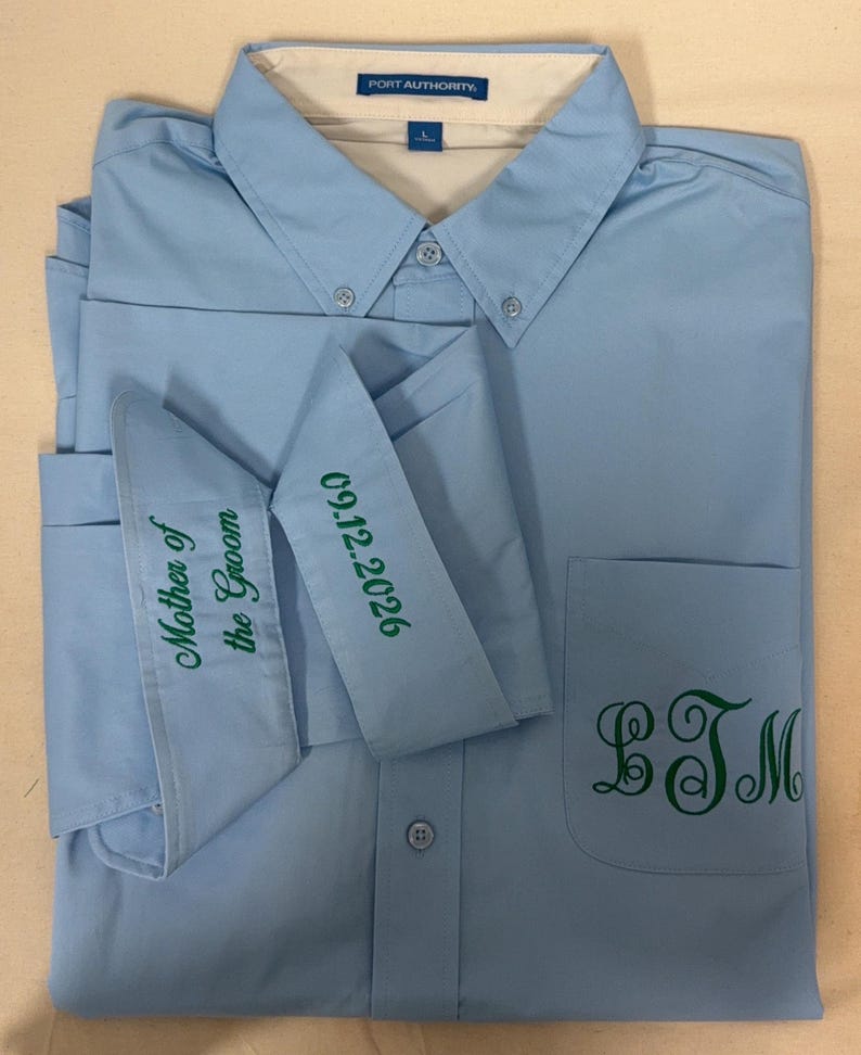 K&ouml;nnte beinhalten: Ein hellblaues Hemd mit Knopfleiste und Port Authority Etikett. Die Manschetten sind gr&uuml;n bestickt mit dem Text Mother of the Groom und 09.12.2026. Ein gr&uuml;nes Monogramm mit den Buchstaben LTM ist auf der Brusttasche aufgestickt.