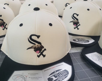 Op de afbeelding: Een stapel witte baseballpetten met zwarte kleppen en een zwart geborduurd logo van een hondenkop met de letter 'S' aan de voorkant. De petten hebben een zwart label aan de zijkant met de tekst 'Flexfit Tech 110 One Ten' en 'Patented Flexfit Technology'.