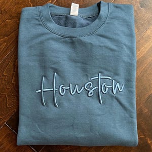 May include: A dark blue sweatshirt with the word "Houston" embroidered in a light blue cursive font.