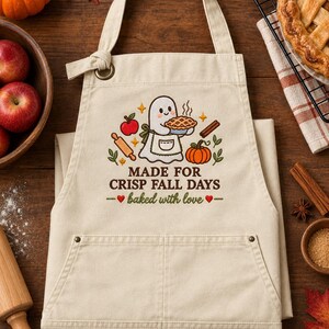 May include: A beige apron featuring a graphic of a ghost wearing an apron and holding a pie, surrounded by an apple, a rolling pin, cinnamon sticks, and a pumpkin. The text reads MADE FOR CRISP FALL DAYS and baked with love with two red hearts.