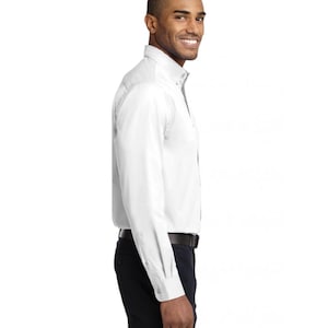 K&ouml;nnte beinhalten: Ein lang&auml;rmeliges wei&szlig;es Hemd mit Button-Down-Kragen und Manschetten, kombiniert mit einer dunklen Hose und einem schwarzen G&uuml;rtel. Das Hemd hat eine durchgehende Knopfleiste auf der Vorderseite und wird in die Hose gesteckt getragen.