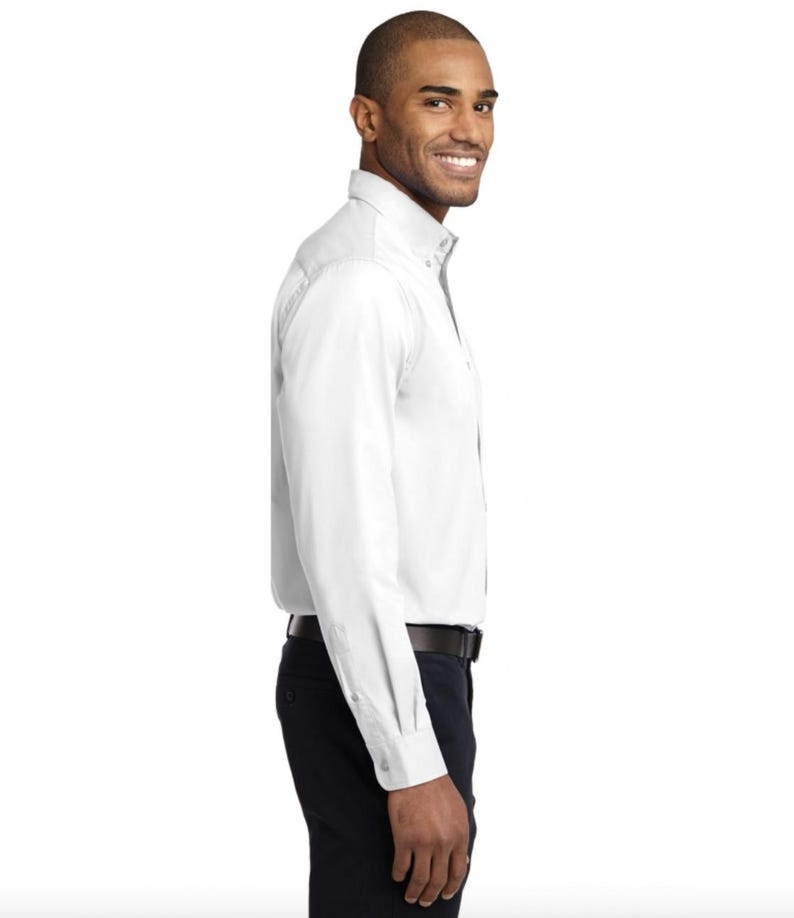 K&ouml;nnte beinhalten: Ein lang&auml;rmeliges wei&szlig;es Hemd mit Button-Down-Kragen und Manschetten, kombiniert mit einer dunklen Hose und einem schwarzen G&uuml;rtel. Das Hemd hat eine durchgehende Knopfleiste auf der Vorderseite und wird in die Hose gesteckt getragen.