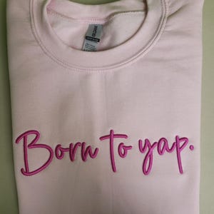 May include: A pink sweatshirt with the phrase "Born to yap." embroidered in pink.