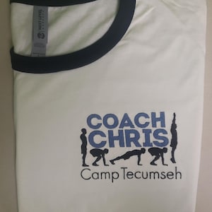 May include: White t-shirt with a black ringer neck. The shirt has an embroidered design that says "Coach Chris" with a blue background and silhouettes of people doing exercises. The text "Camp Tecumseh" is below the design.