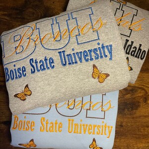 May include: Two gray and blue sweatshirts with the text "Boise State University" and "Broncos" embroidered in orange and blue. The sweatshirts have a butterfly design embroidered on them.