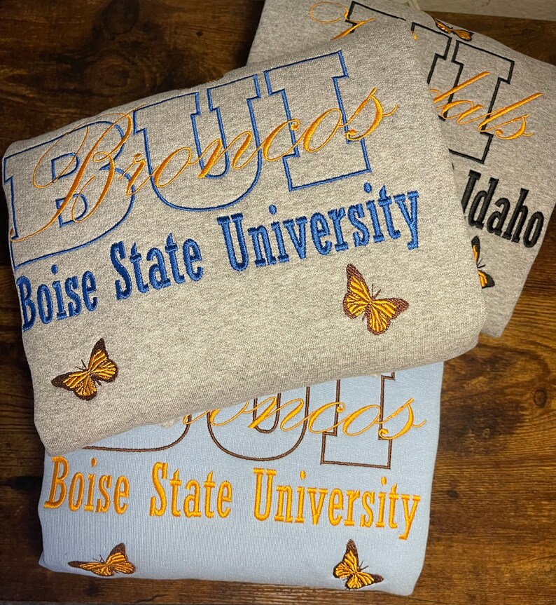 May include: Two gray and blue sweatshirts with the text "Boise State University" and "Broncos" embroidered in orange and blue. The sweatshirts have a butterfly design embroidered on them.