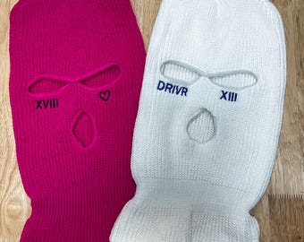 以下が含まれることがあります： 鮮やかなピンクと白のニット製バラクラバ2点。ピンクのバラクラバには黒い刺繍で「XVIII」と小さなハートのシンボルが施されています。白いバラクラバには青い刺繍で「DRIVR」とローマ数字の「XIII」が施されています。