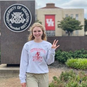 May include: A woman wearing a gray sweatshirt with the words "University of Houston" and a red mascot logo. She is standing in front of a building with a red "UH" logo.