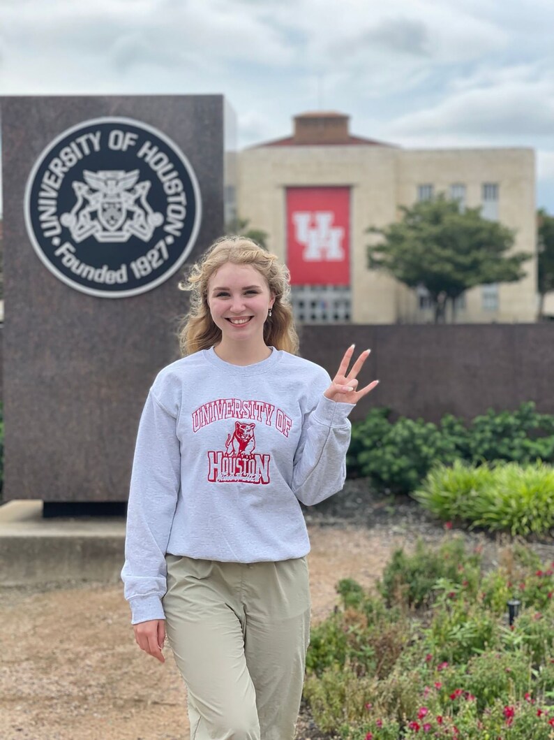 May include: A woman wearing a gray sweatshirt with the words "University of Houston" and a red mascot logo. She is standing in front of a building with a red "UH" logo.