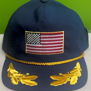 May include: Navy blue baseball cap featuring an embroidered American flag patch. The cap has a gold rope detail along the brim and gold leaf accents. A classic and patriotic accessory.