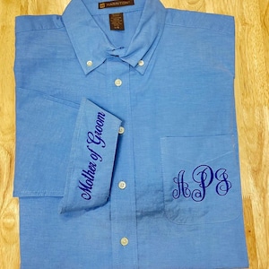 K&ouml;nnte beinhalten: Ein hellblaues Hemd mit kurzen &Auml;rmeln, Kragen und einer Brusttasche. Das Hemd ist mit einer dunkelblauen Stickerei am &Auml;rmelaufschlag mit der Aufschrift Mother of Groom und einem dunkelblauen Monogramm mit den Buchstaben APS versehen.