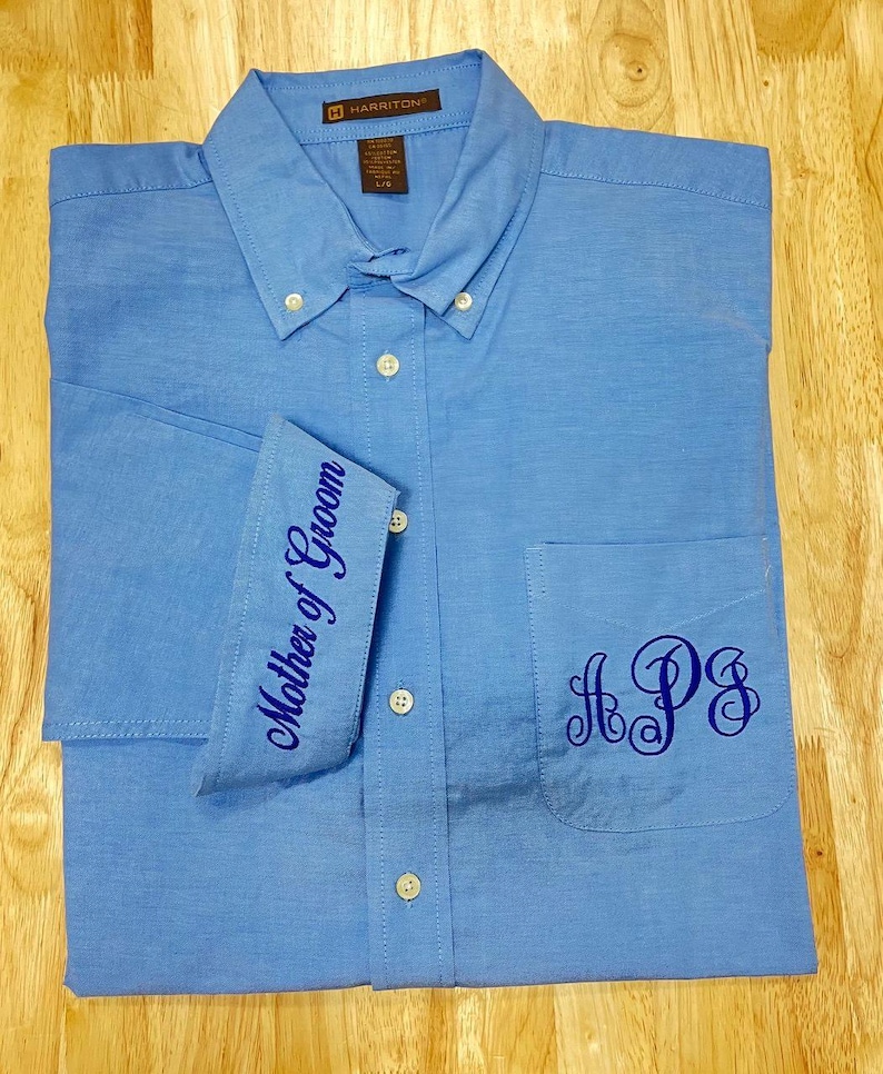 K&ouml;nnte beinhalten: Ein hellblaues Hemd mit kurzen &Auml;rmeln, Kragen und einer Brusttasche. Das Hemd ist mit einer dunkelblauen Stickerei am &Auml;rmelaufschlag mit der Aufschrift Mother of Groom und einem dunkelblauen Monogramm mit den Buchstaben APS versehen.