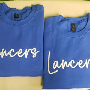 May include: Two royal blue t-shirts with white embroidered text that reads "Lancers".