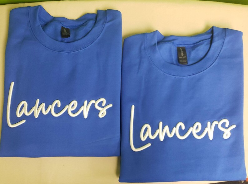 May include: Two royal blue t-shirts with white embroidered text that reads "Lancers".