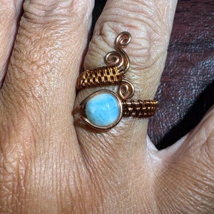 May include: A ring featuring a round, light blue stone set in a copper wire frame. The copper wire is intricately wrapped around the band and features decorative spiral accents at the top, creating a textured, metallic appearance around the stone.