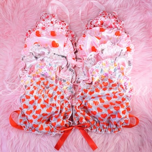 May include: A set of layered lingerie tops with a pink and red heart print. The tops are made of satin and have a ruffled trim. The tops are tied at the neck with a red ribbon.