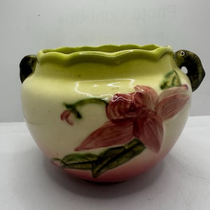 May include: A vintage ceramic planter with a cream-colored base and a scalloped, light green rim. The planter features a large, red and pink floral design and two small handles. A charming piece for displaying plants.