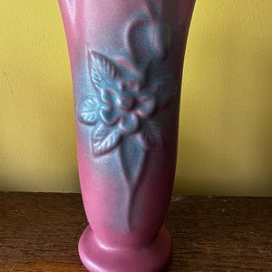 Puede incluir: Un jarrón alto y vintage en tonos rosa y azul. El jarrón de cerámica presenta un diseño floral en relieve en la parte delantera. El jarrón tiene una parte superior acampanada y una base redonda. El jarrón está sobre una superficie de madera.