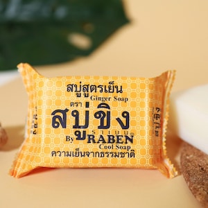 May include: A yellow and orange bar of soap wrapped in a foil packet. The packet has Thai text that reads "สบู่ขิง" which translates to "Ginger Soap" and "By URABEN Cool Soap". The text also says "ความเย็นจากธรรมชาติ" which translates to "Natural coolness".